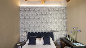A bedroom with a patterned wallpaper, a bed with navy pillows, a bedside table with a book and a vase, and a desk with a chair and a vase.