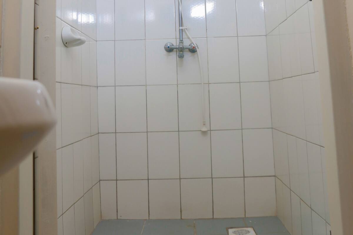 A small, tiled shower room with a blue mat on the floor, a handheld showerhead, and a wall-mounted light fixture.