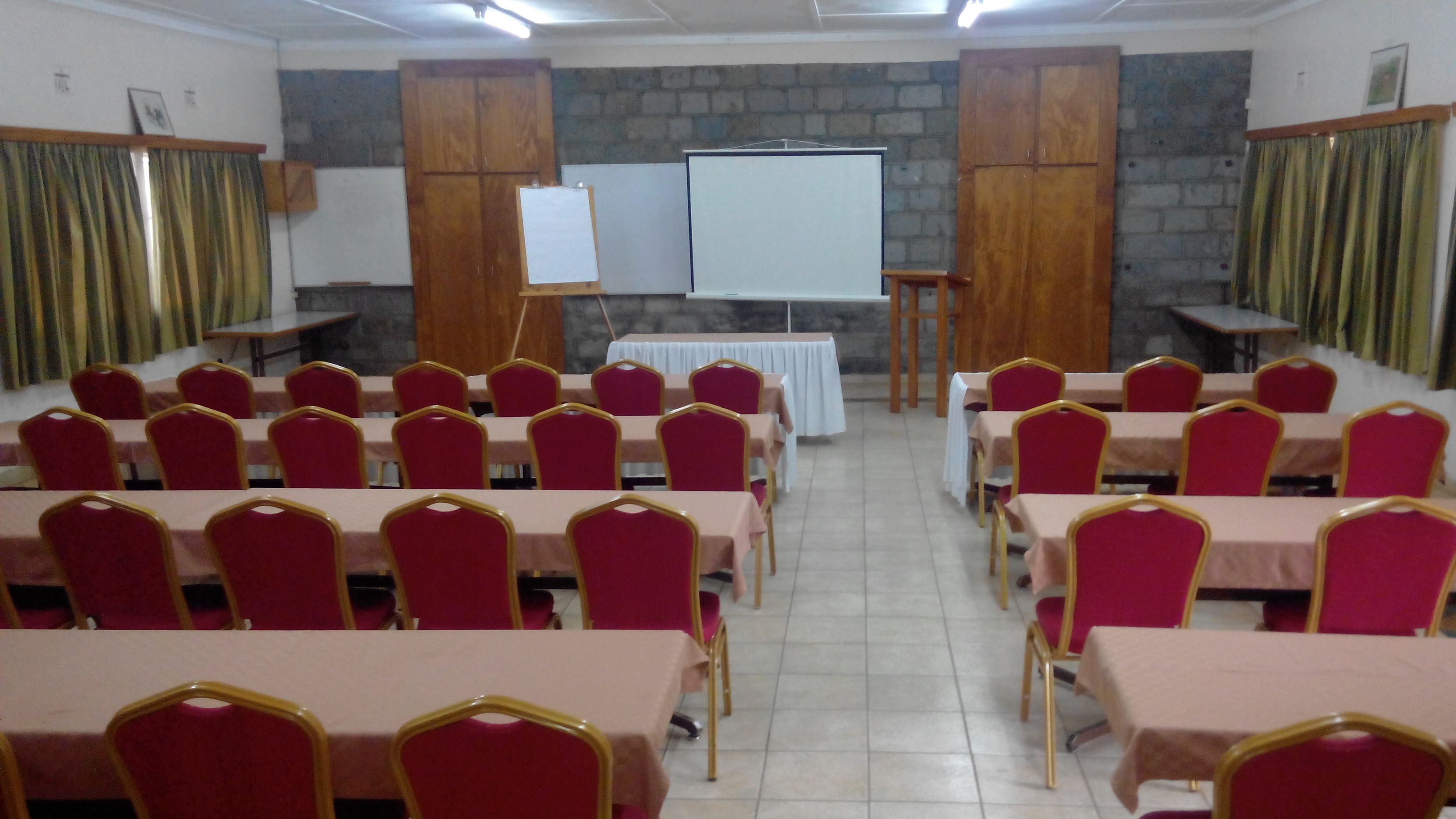 Un salón de conferencias con filas de sillas rojas y mesas, una pizarra y una pantalla de proyector.