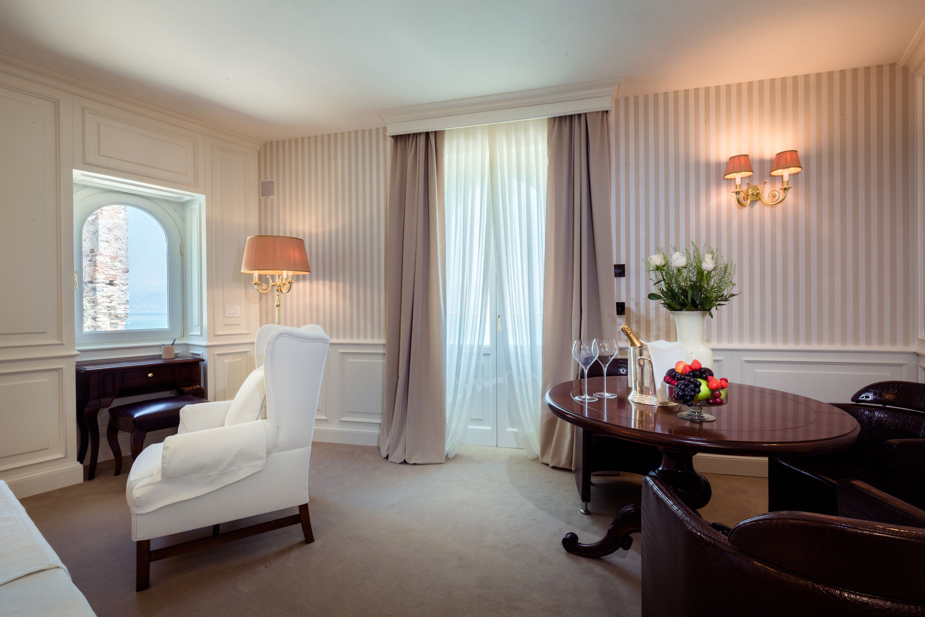 A hotel room with a white armchair, a wooden table with a vase of flowers, a bottle of champagne, and a fruit bowl.