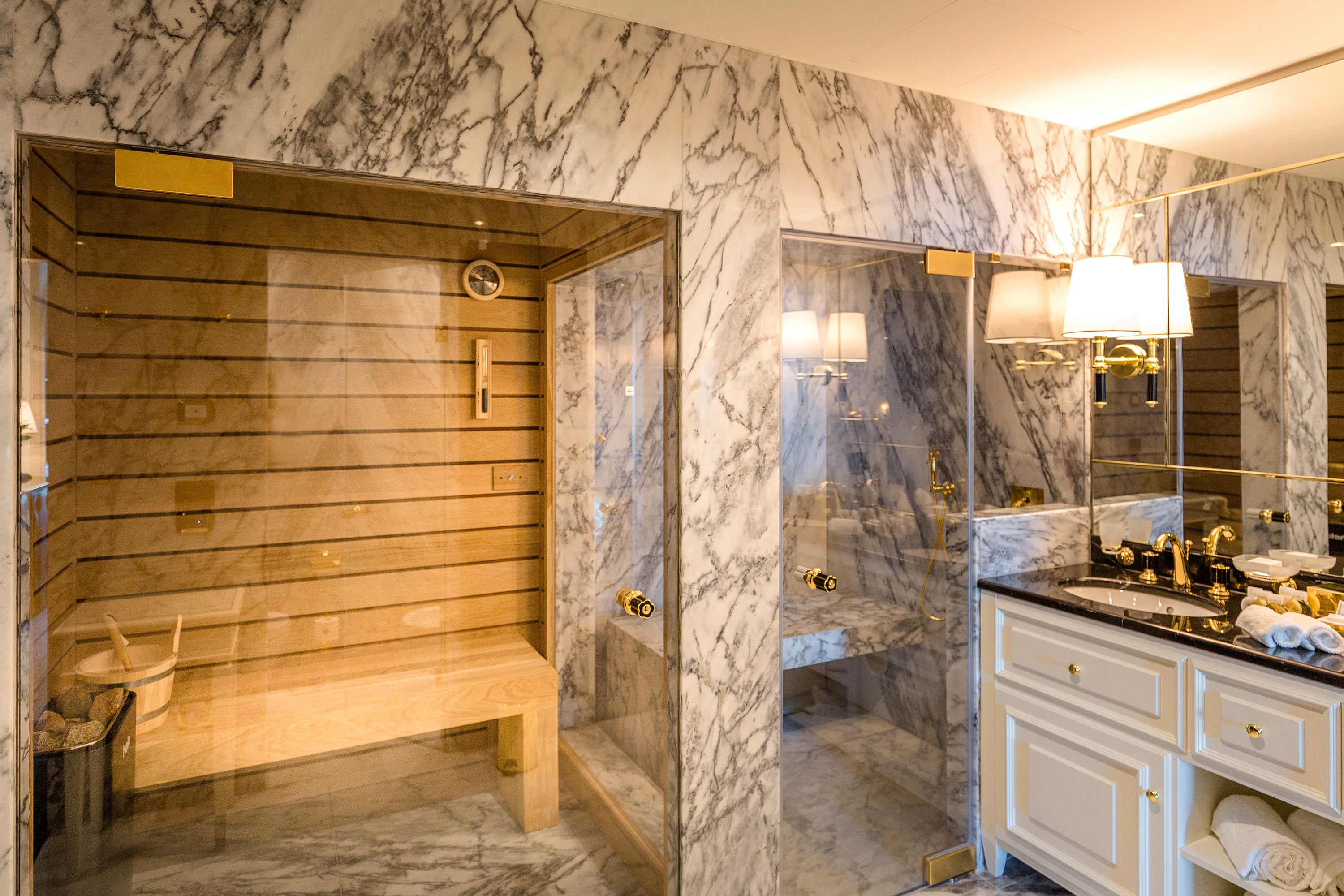 Een moderne badkamer met een houten sauna, marmeren wanden en een wastafel met gouden details.