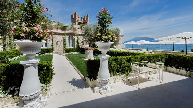 A courtyard with white urns, a white outdoor seating area, and a view of a castle-like structure. - Relais La Speranzina (Sirmione)