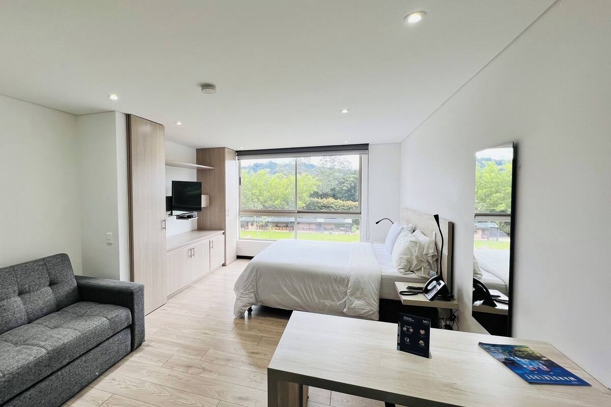 A modern bedroom with a large bed, a grey sofa, a TV, and a window with a view of greenery.