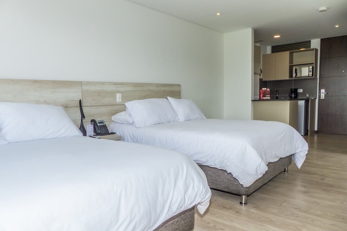 A hotel room with two beds, a bedside phone, and a kitchenette in the background.