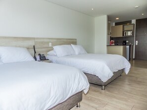 A hotel room with two beds, a bedside phone, and a kitchenette in the background. - MG Hotels & Suites (Rionegro)