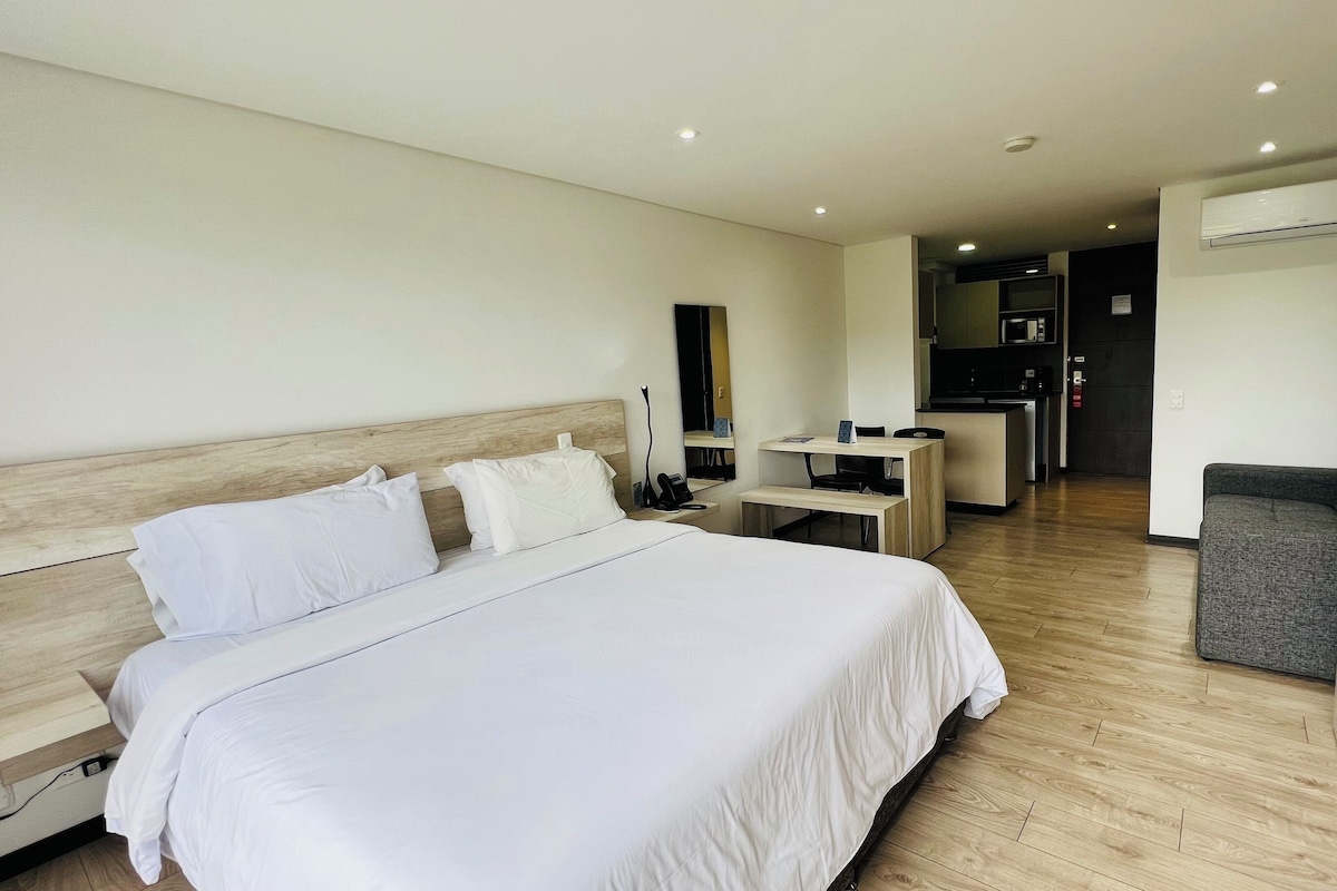 A hotel room with a large bed, a desk, a chair, a small table, and a kitchenette in the background.