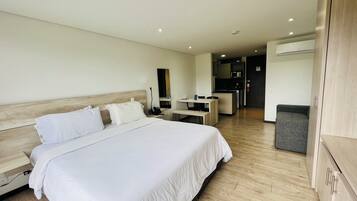 Habitación de hotel con una cama grande, un escritorio, una silla, una mesita y una kitchenette al fondo.