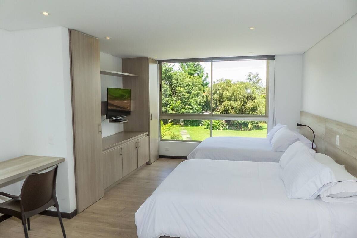 A modern hotel room with a desk, two beds, a TV, and a large window with a view of greenery.