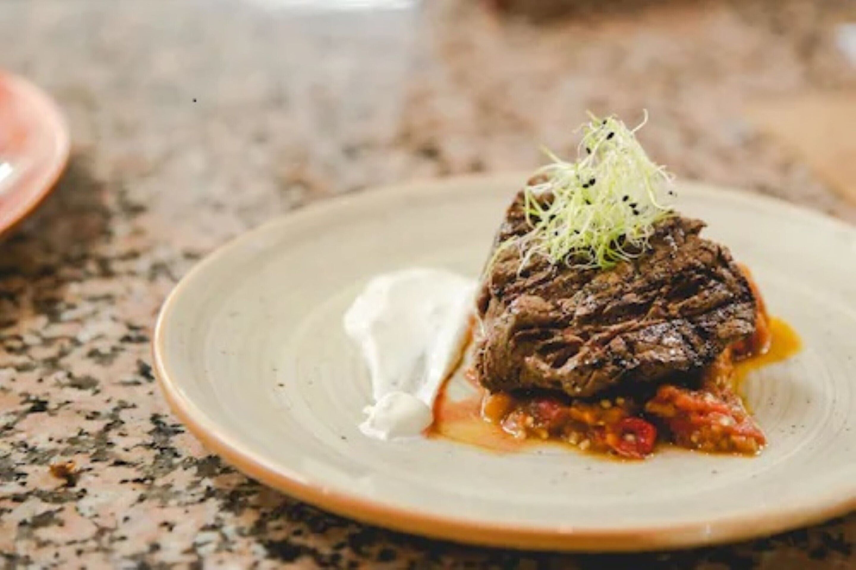 A plate with a meat dish, possibly beef, topped with a garnish, served with a side of sauce.