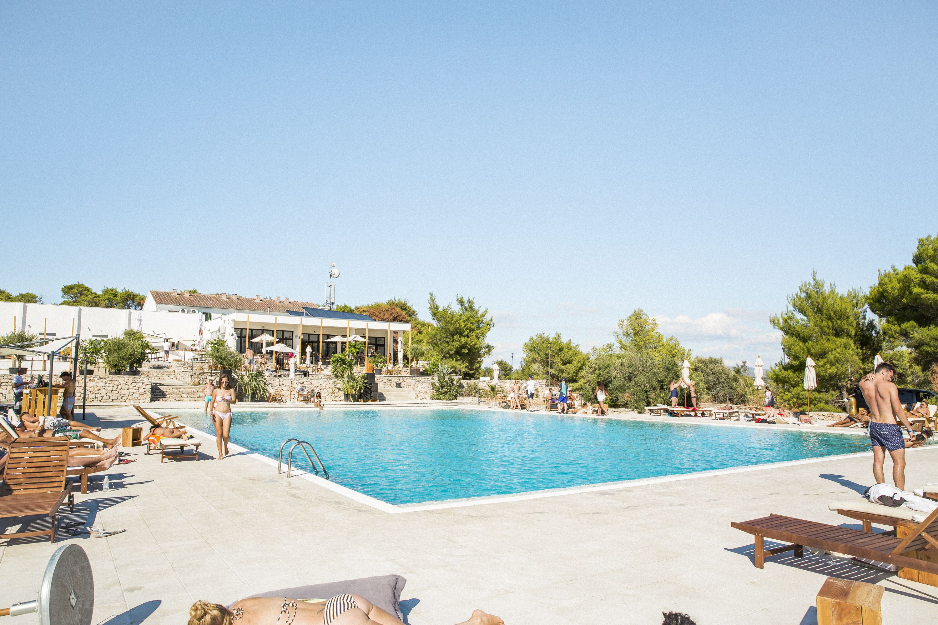 A swimming pool with lounge chairs and umbrellas, surrounded by trees and a building with a tiled roof.