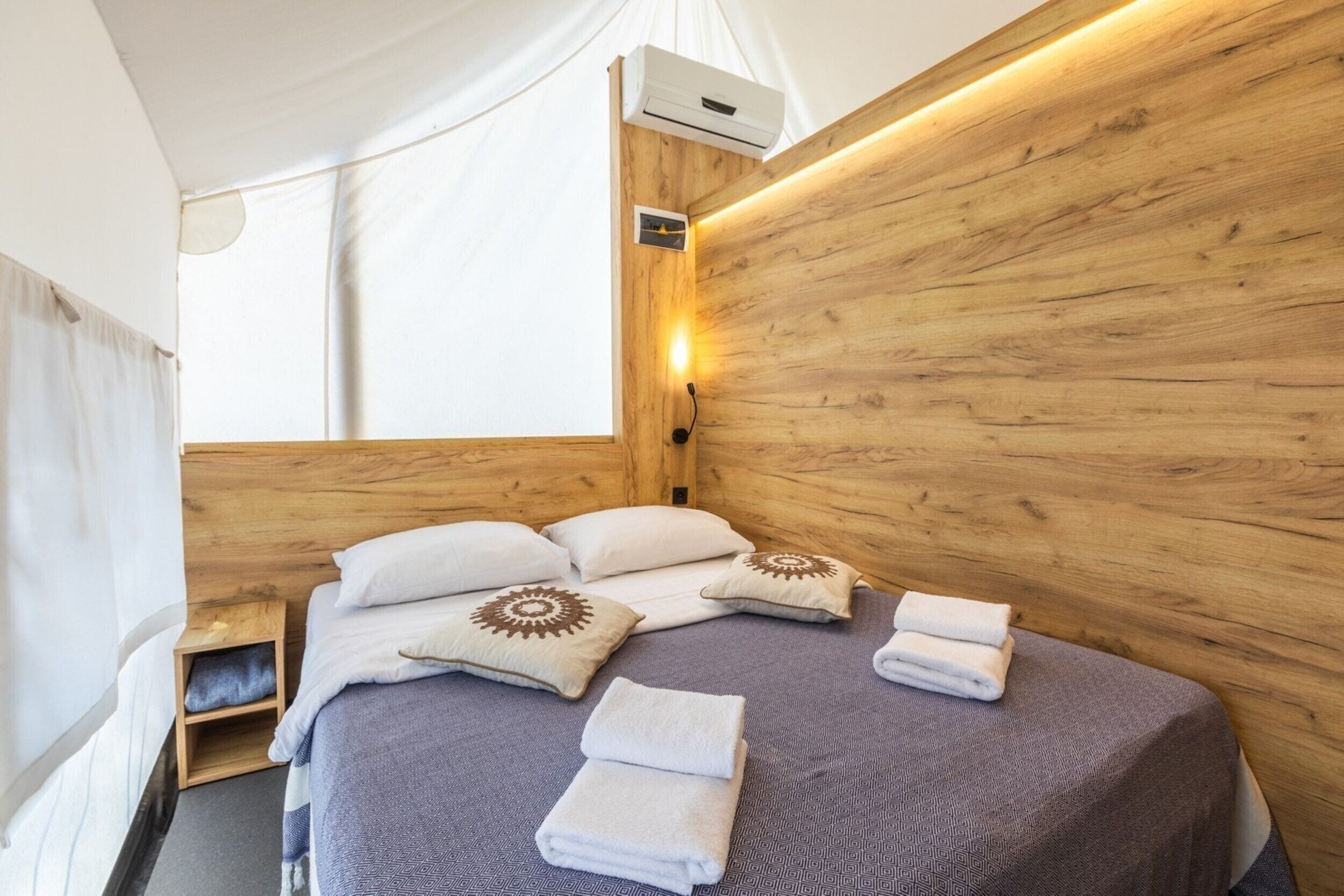 Ein gemütliches Schlafzimmer mit einem hölzernen Kopfteil, einem Bett mit weißen und gemusterten Kissen sowie einem ordentlich bezogenen Bett mit gefalteten Handtüchern.