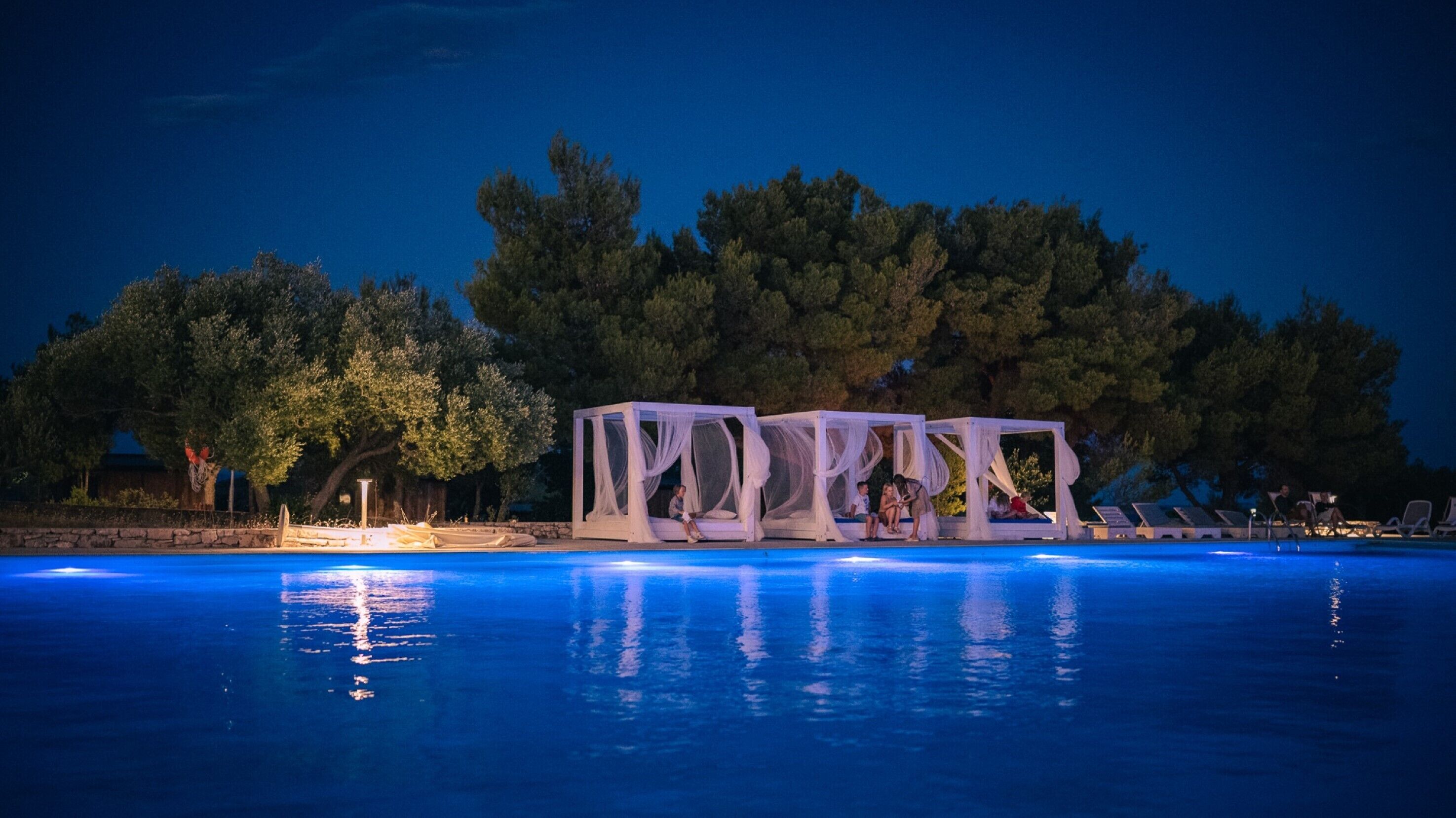 Un espace aménagé au bord de la piscine, offrant une vue dégagée sur l’eau, avec une rangée de chaises longues et une rangée de coins salon couverts de toiles blanches.