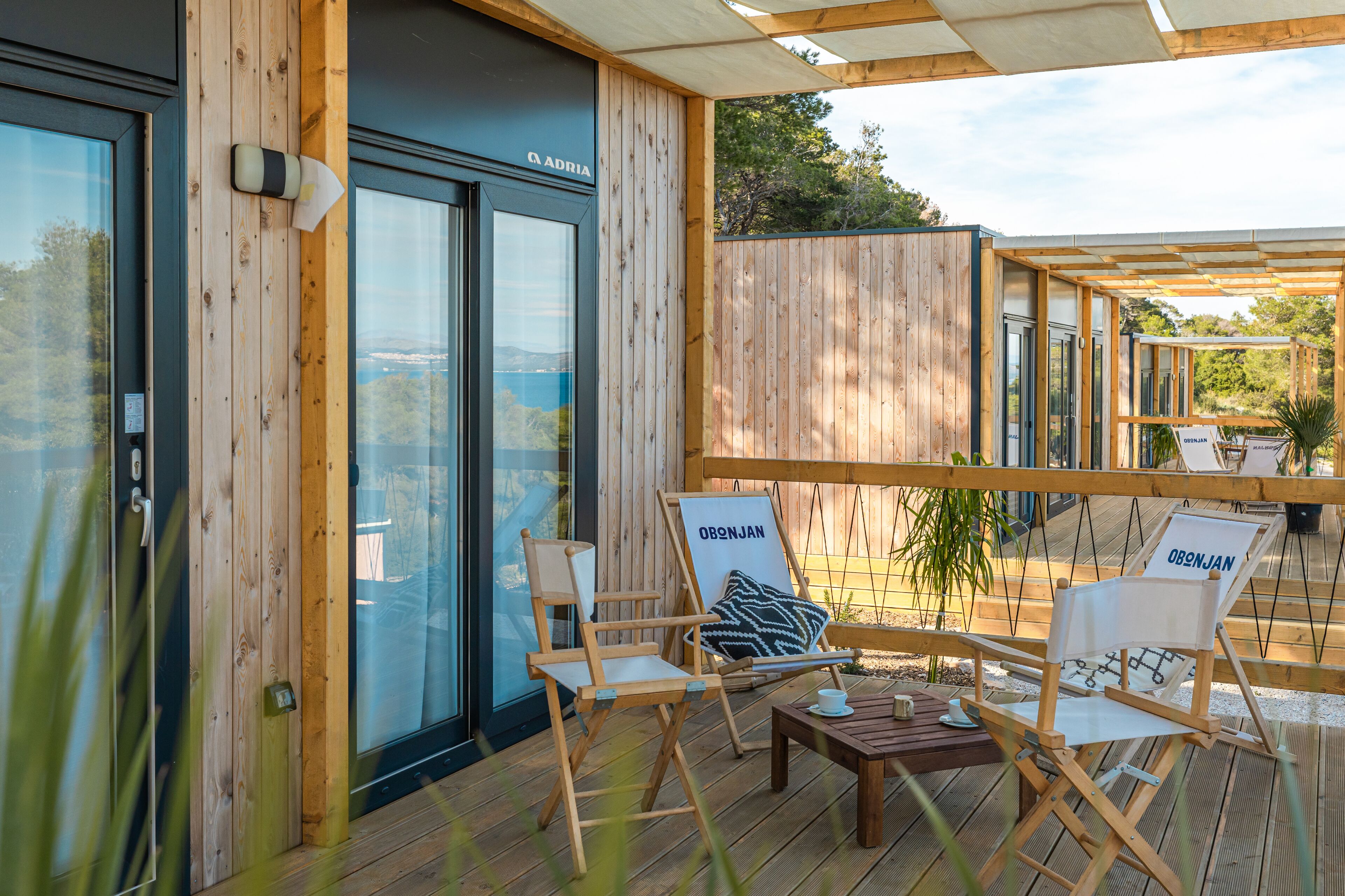Une terrasse en bois avec deux chaises pliantes et une table, une porte vitrée et une vue sur l’extérieur.