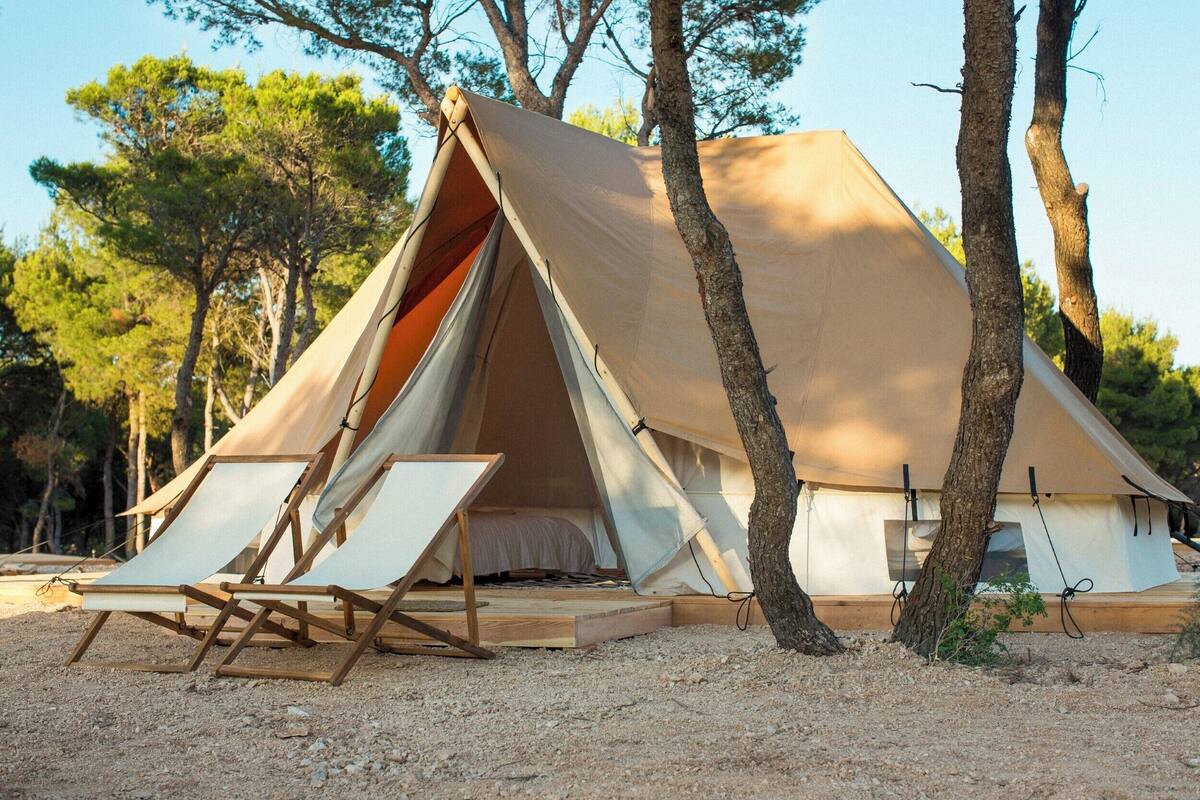 A large, A-frame tent with a wooden platform and a bed inside, surrounded by trees.