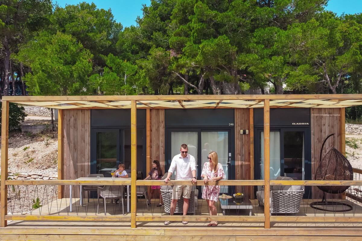 A modern wooden cabin with a glass facade, a dining area with outdoor seating, and a stone wall in the background.