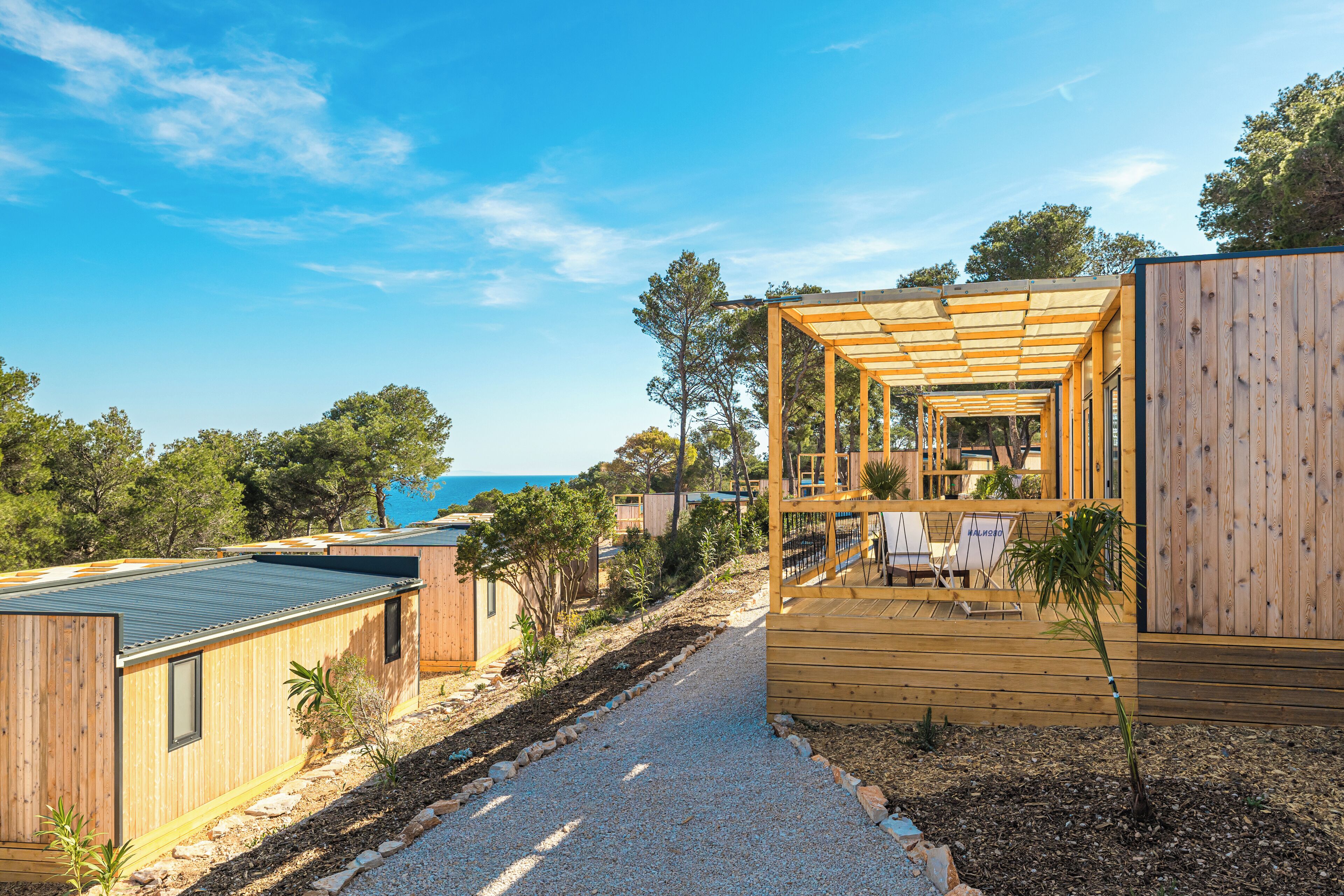Una cabaña de madera moderna con terraza y zona de estar exterior, rodeada de árboles y un cielo despejado.