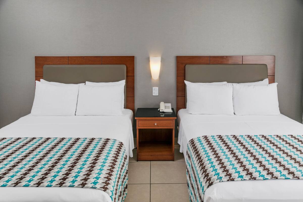 Een hotelkamer met twee bedden, elk met witte beddengoed en een gestreept plaid. Tussen de bedden staat een nachtkastje met een telefoon en een klein lade.