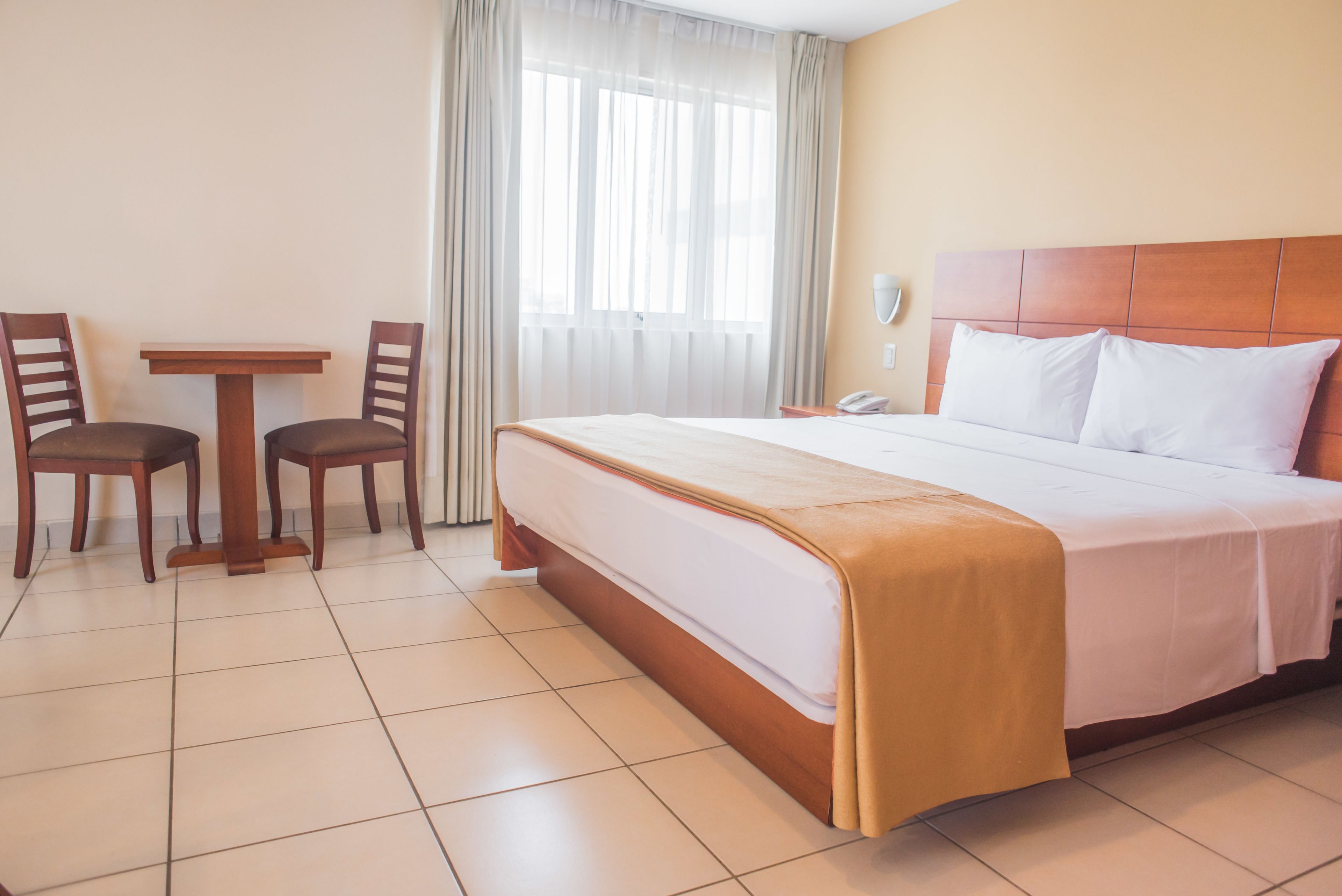 Habitación de hotel con una cama grande, una mesita pequeña, dos sillas, un teléfono y una ventana con cortinas.
