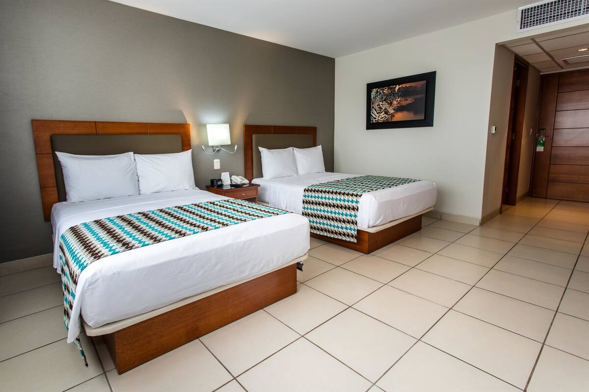 Een hotelkamer met twee bedden, elk voorzien van een houten hoofdeinde, wit beddengoed en een gestreept deken. Er is een nachtkastje met een lamp en een telefoon, en een lijstje aan de muur.