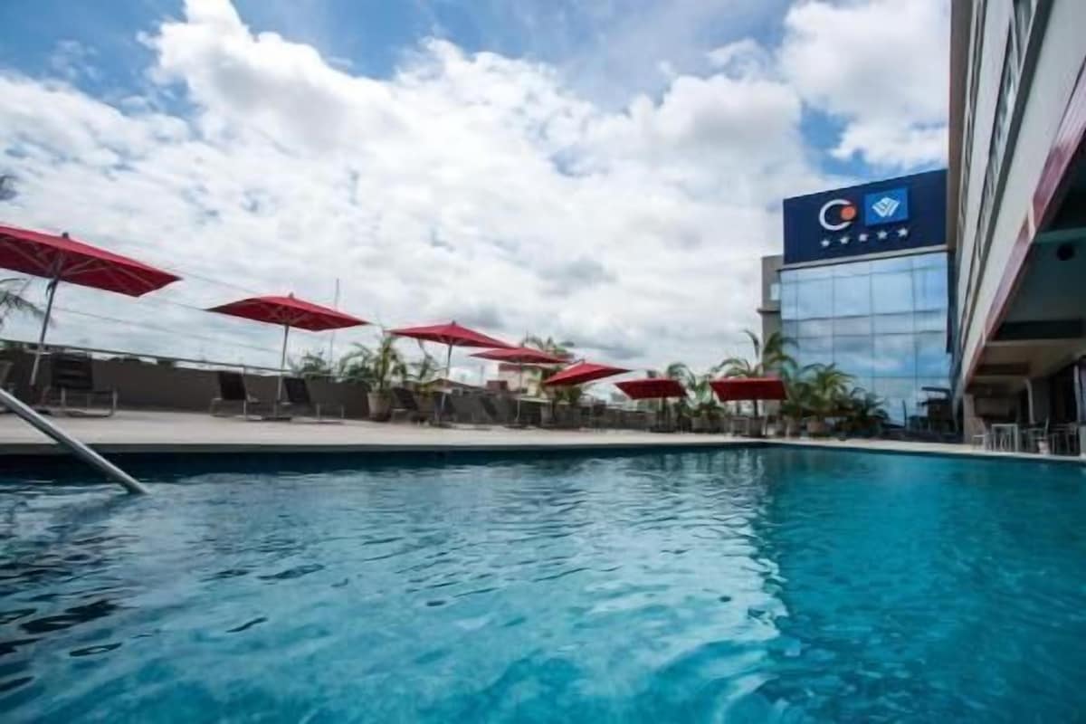 Una piscina con sombrillas rojas, sillas de descanso y un edificio de hotel con un logotipo.