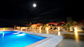 Área de piscina nocturna con sillones de descanso y sombrillas, bajo un cielo estrellado con luna visible.