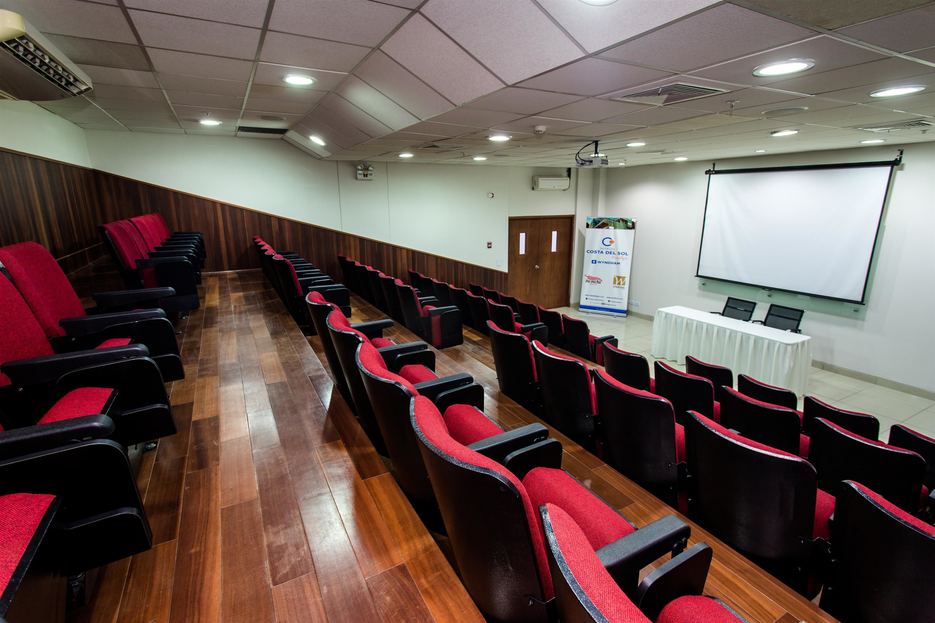 A conference room with rows of black chairs with red cushions, a wooden floor, a white projection screen, and a table with a white tablecloth.