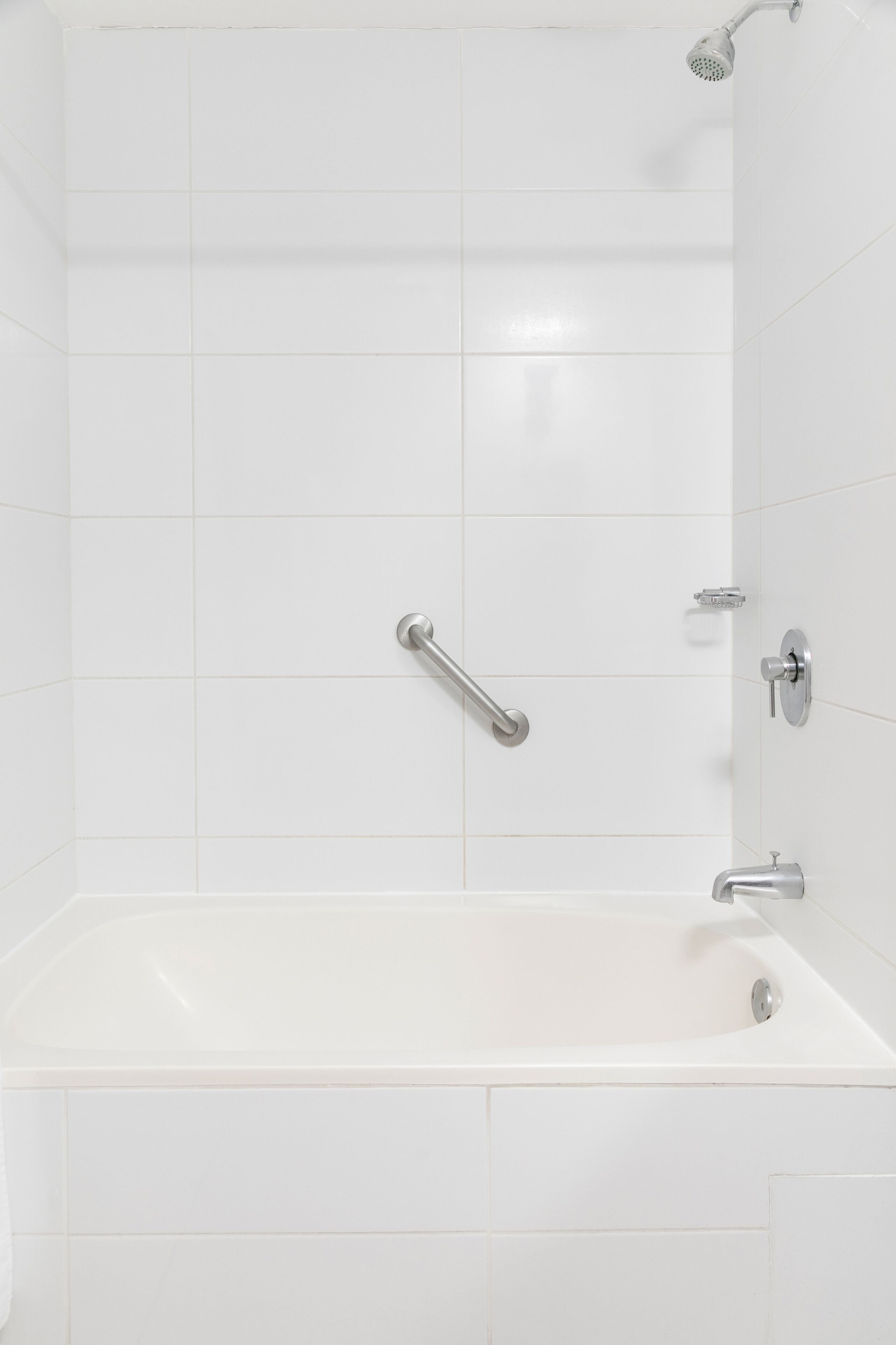 A modern bathroom with white tiles, a bathtub, a handheld showerhead, and a grab bar.