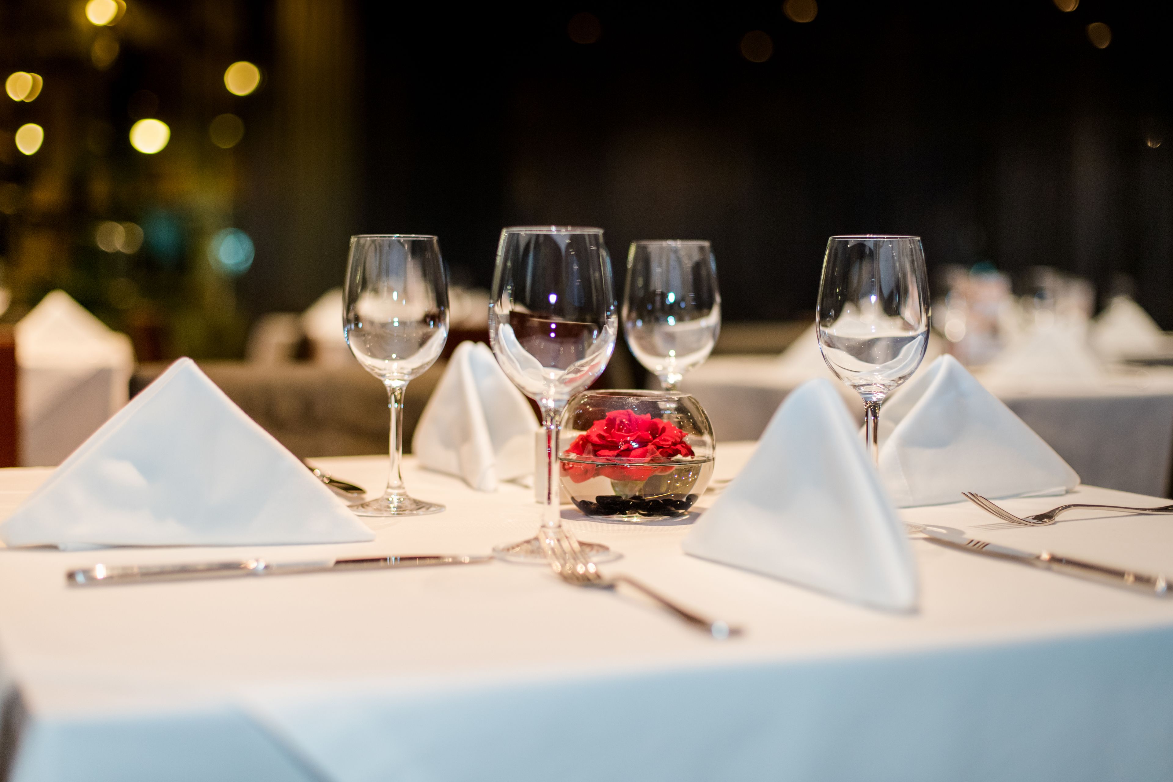 Una mesa lista para comer con copas de vino, un cuenco pequeño con flores rojas y servilletas blancas.