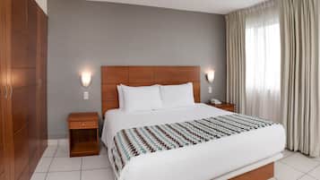 A hotel room with a large bed, wooden headboard, two bedside tables with lamps, a wardrobe, and a window with curtains.