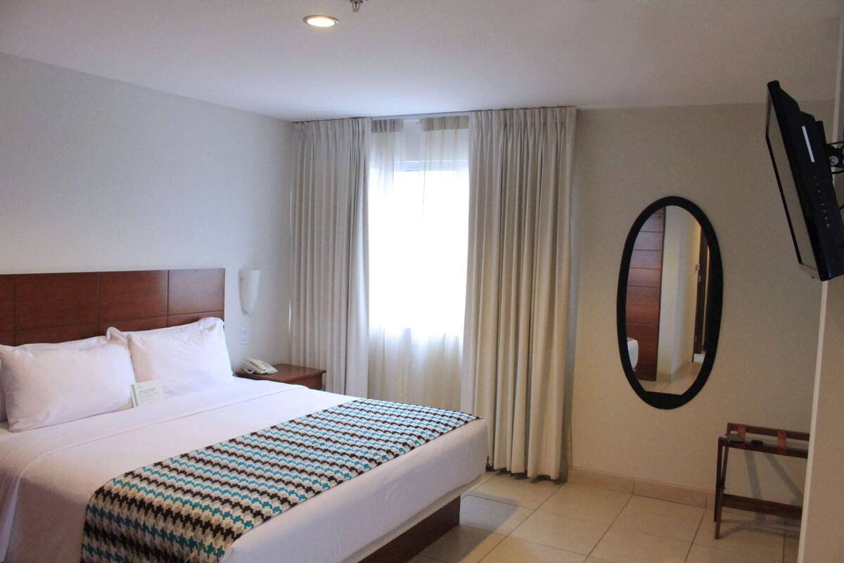 Een hotelkamer met een groot bed, een nachtkastje met een telefoon, een spiegel aan de muur en een aan de muur bevestigde televisie.