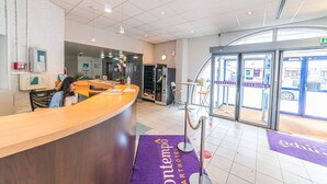 A hotel reception area with a wooden counter, a vending machine, and a purple carpet with the hotel name. - Montempô Apparthôtel Evry (Évry-Courcouronnes)