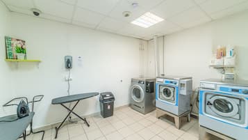 A laundry room with two washing machines, an ironing station, and a trash bin.