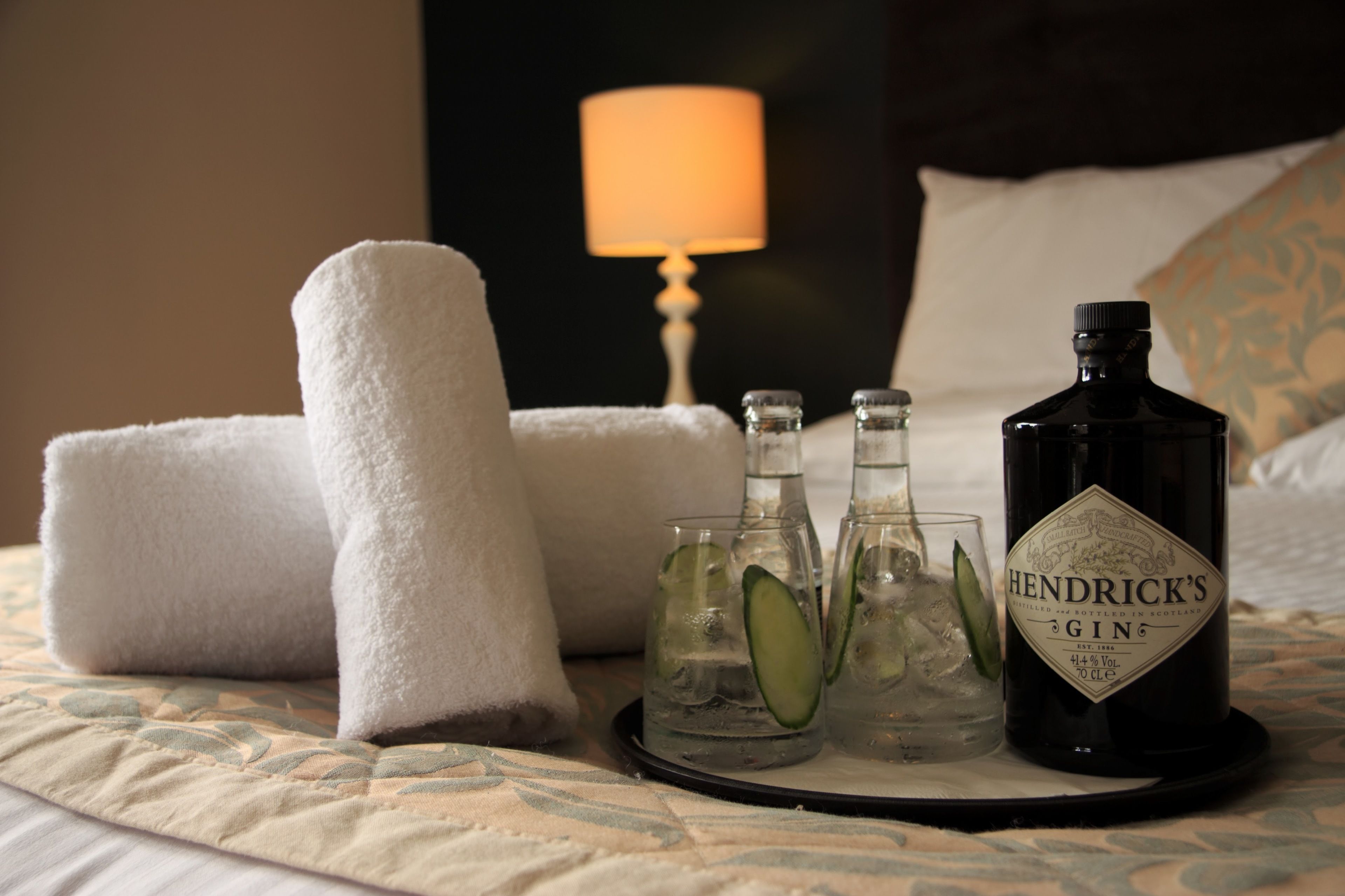 Una cama con una bandeja que contiene una botella de ginebra Hendrick's, dos vasos con hielo y rodajas de pepino, y dos toallas dobladas.