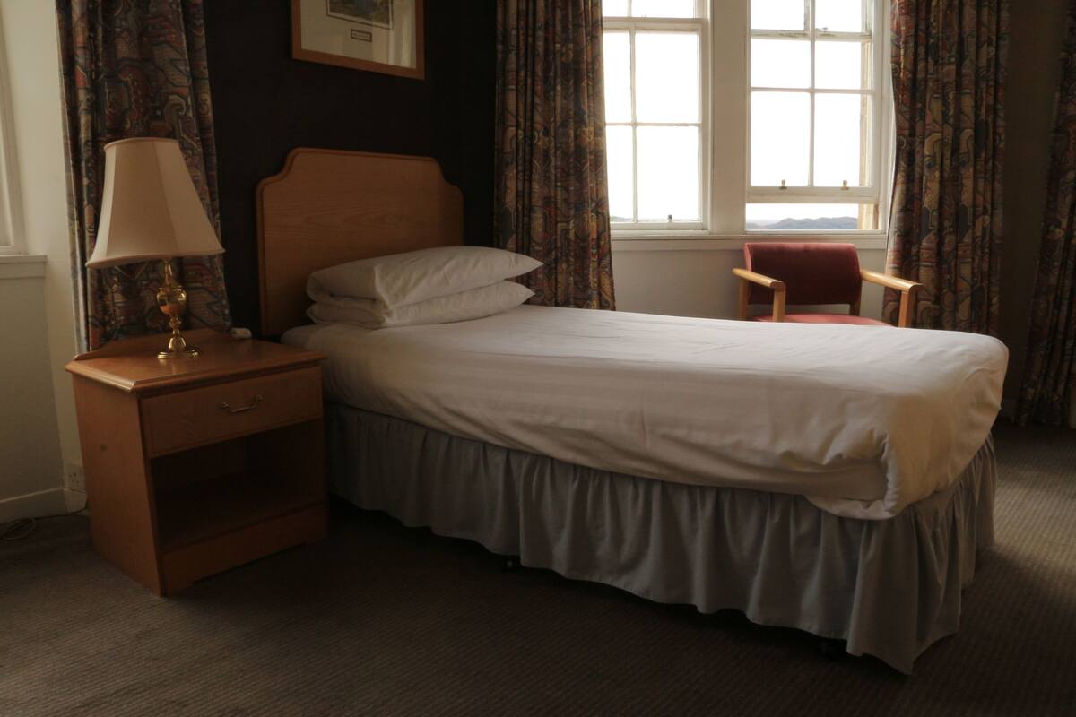 Een net opgemaakt bed met een houten hoofdbord, een nachtkastje met een lamp en een stoel bij het raam.