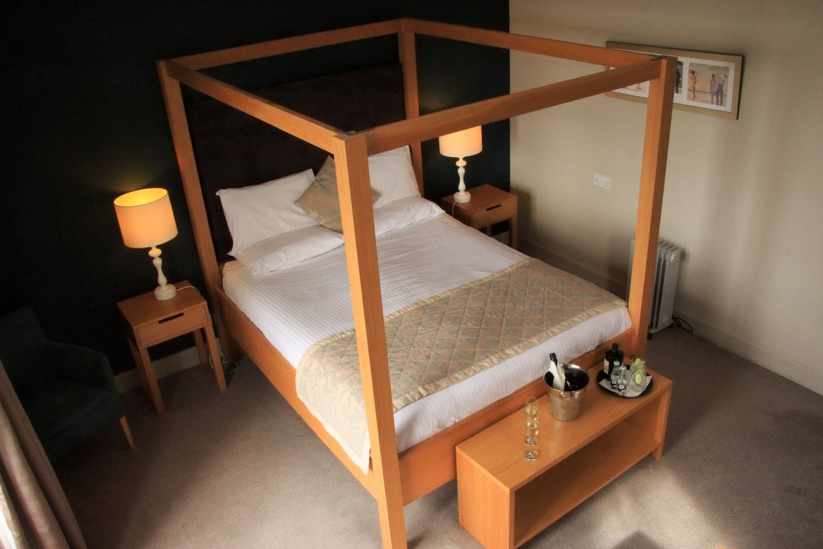 Een hemelbed met houten frame, twee nachtkastjes met lampen, een klein nachtkastje met een fles en glazen, en een ingelijst beeld aan de muur.