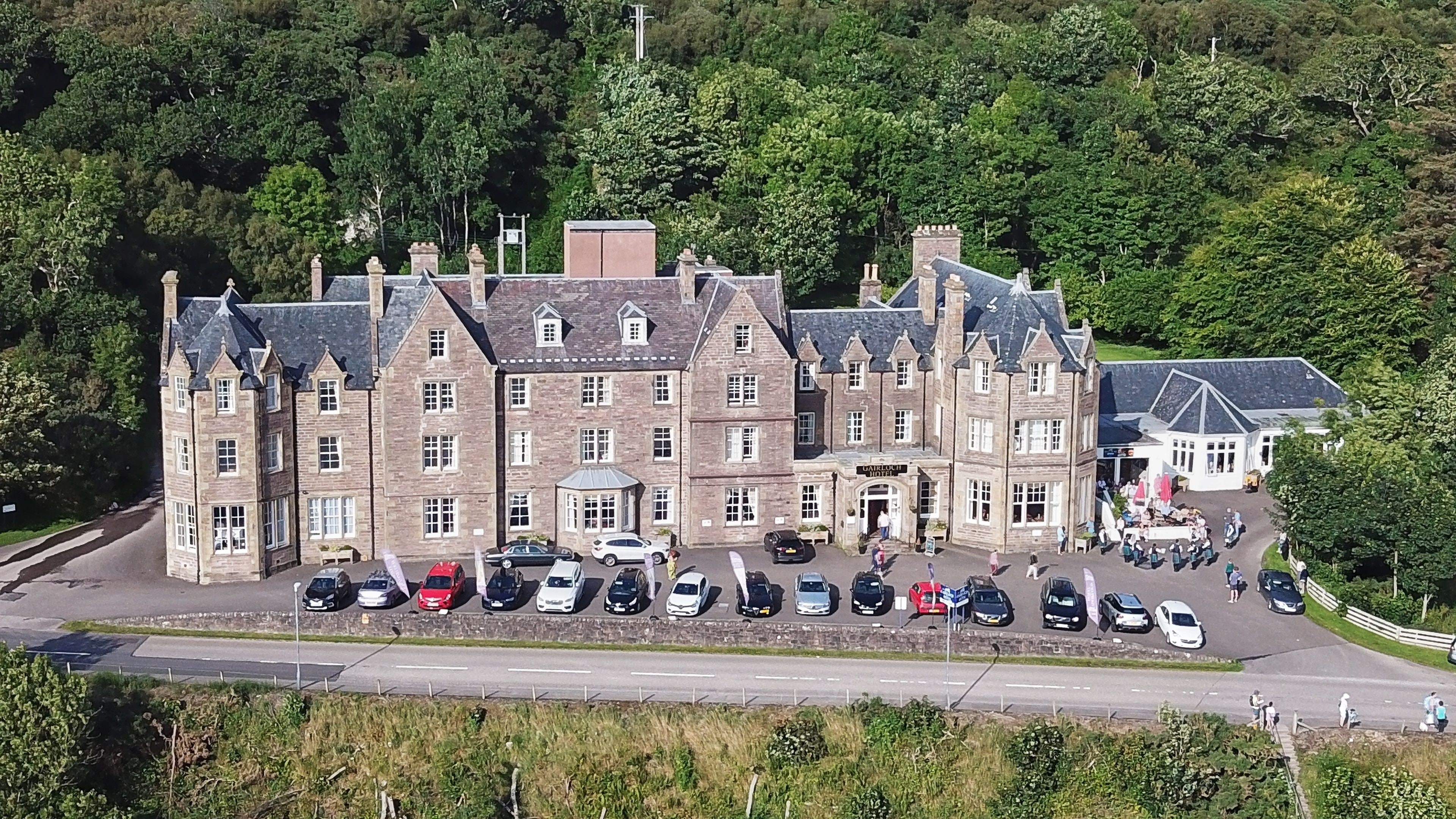 A large, historic building with multiple stories and a prominent entrance. There are several cars parked in front, and people are visible on the ground floor.