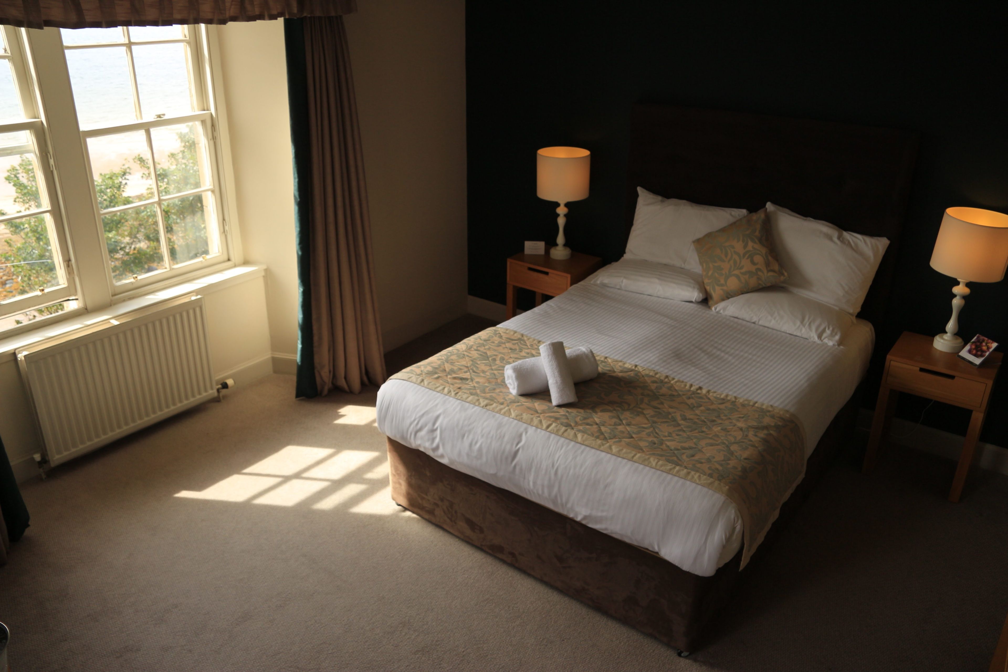 A neatly made bed with a patterned bedspread, two bedside lamps, and a window with curtains.