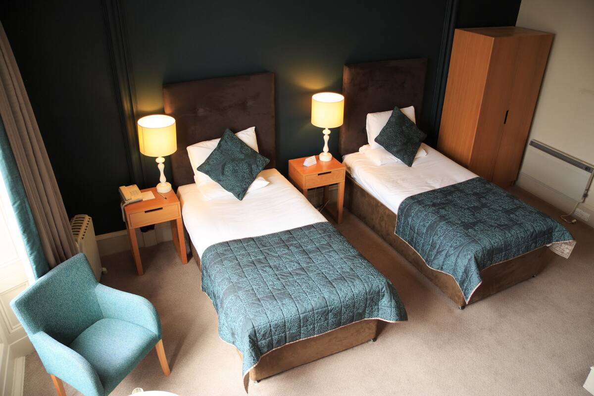 Een hotelkamer met twee bedden, een stoel, een kledingkast en een nachtkastje met een lamp.