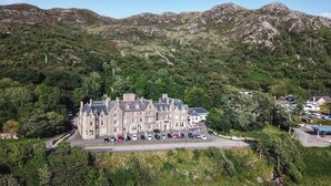 Un edificio grande e histórico con varias secciones, rodeado de abundante vegetación y montañas.