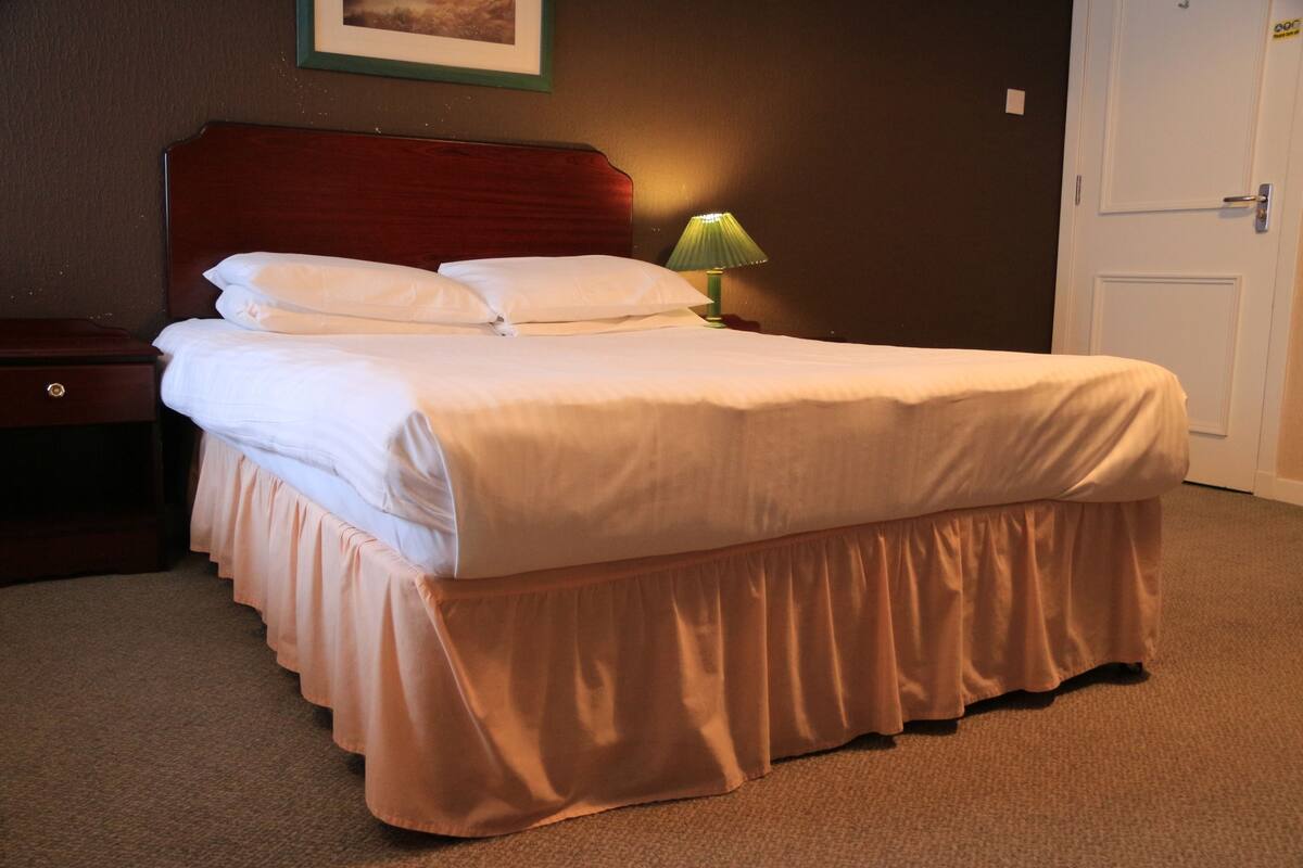 Een net opgemaakt bed met een houten hoofdbord, een groene bedlamp en een gesloten deur in een hotelkamer.
