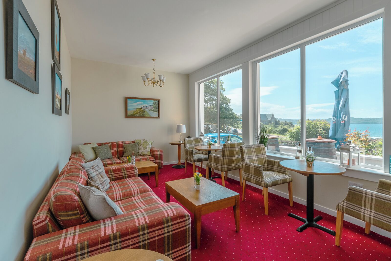 A living room with a red carpet, plaid sofa, and chairs, large windows with a view of the sea, and a chandelier.