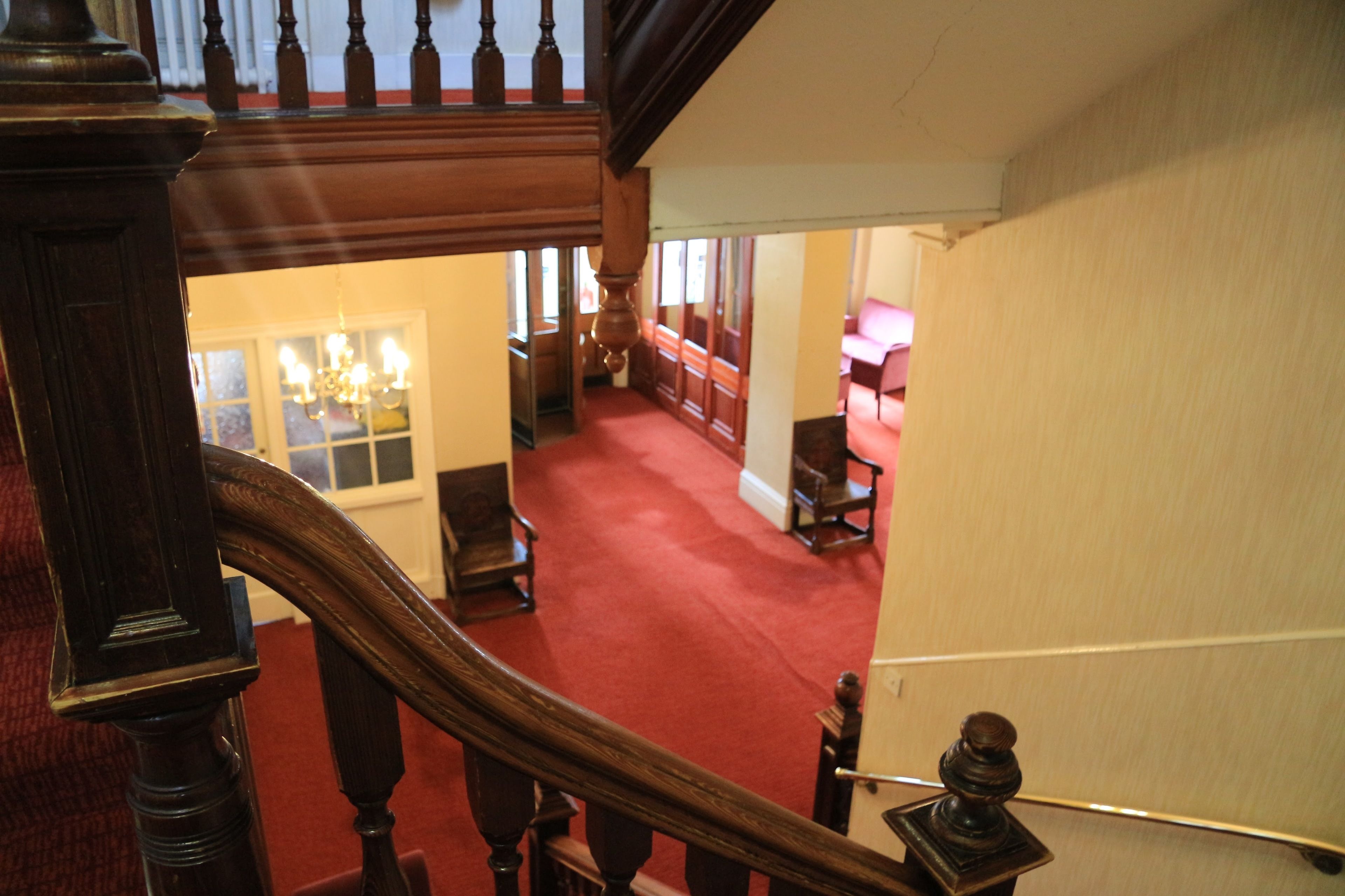 A staircase with wooden balusters leading to a room with a red carpet, a chandelier, and wooden furniture.