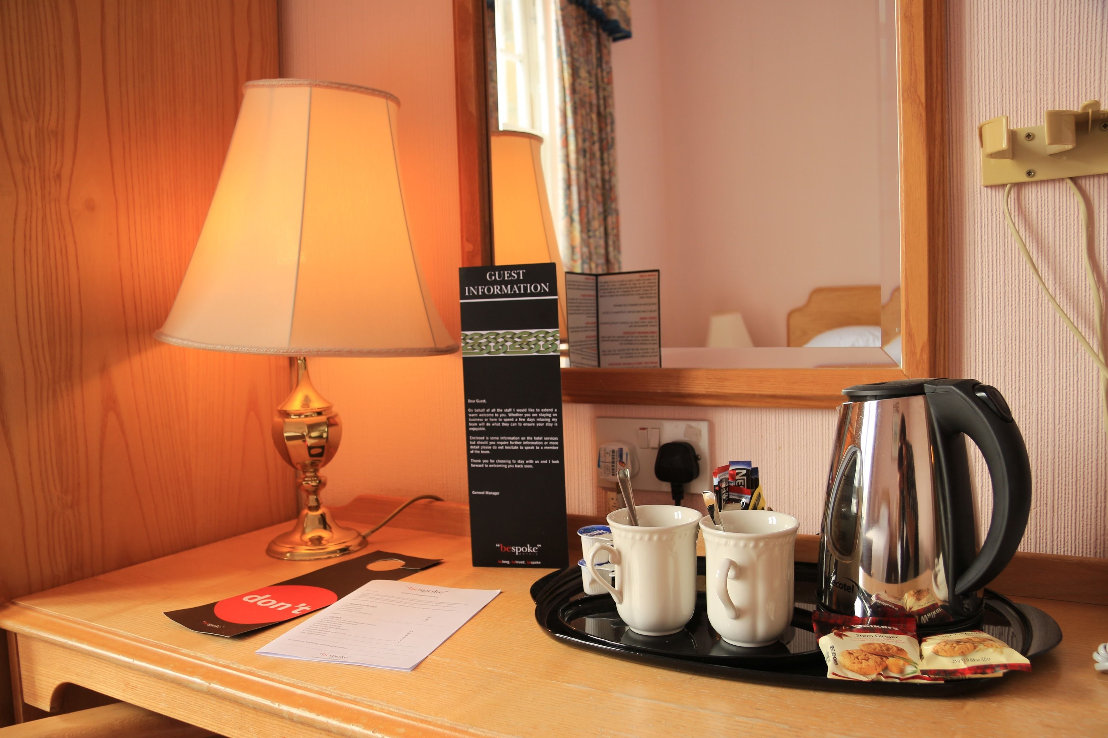 A hotel room desk with a lamp, a tray with mugs, a kettle, and a guest information card.