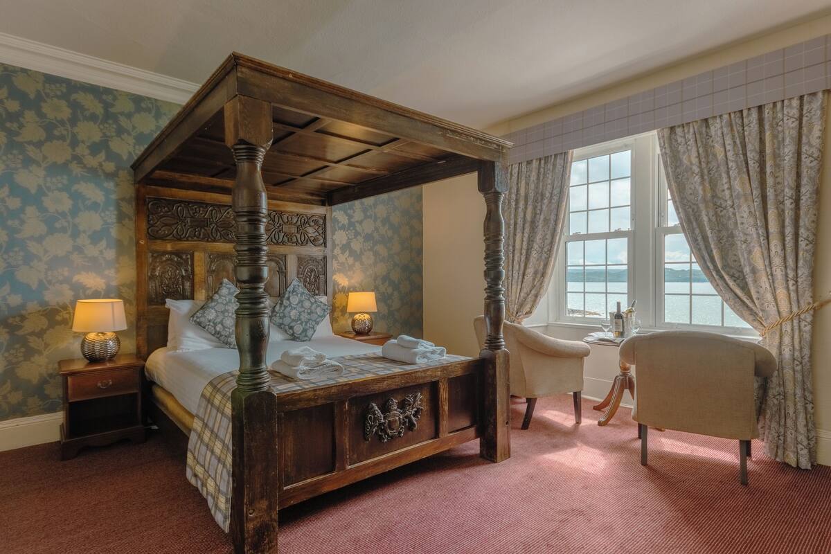 Een hemelbed met een houten hoofdbord, twee nachtkastjes met lampen, een stoel en uitzicht op zee door een raam met gordijnen.