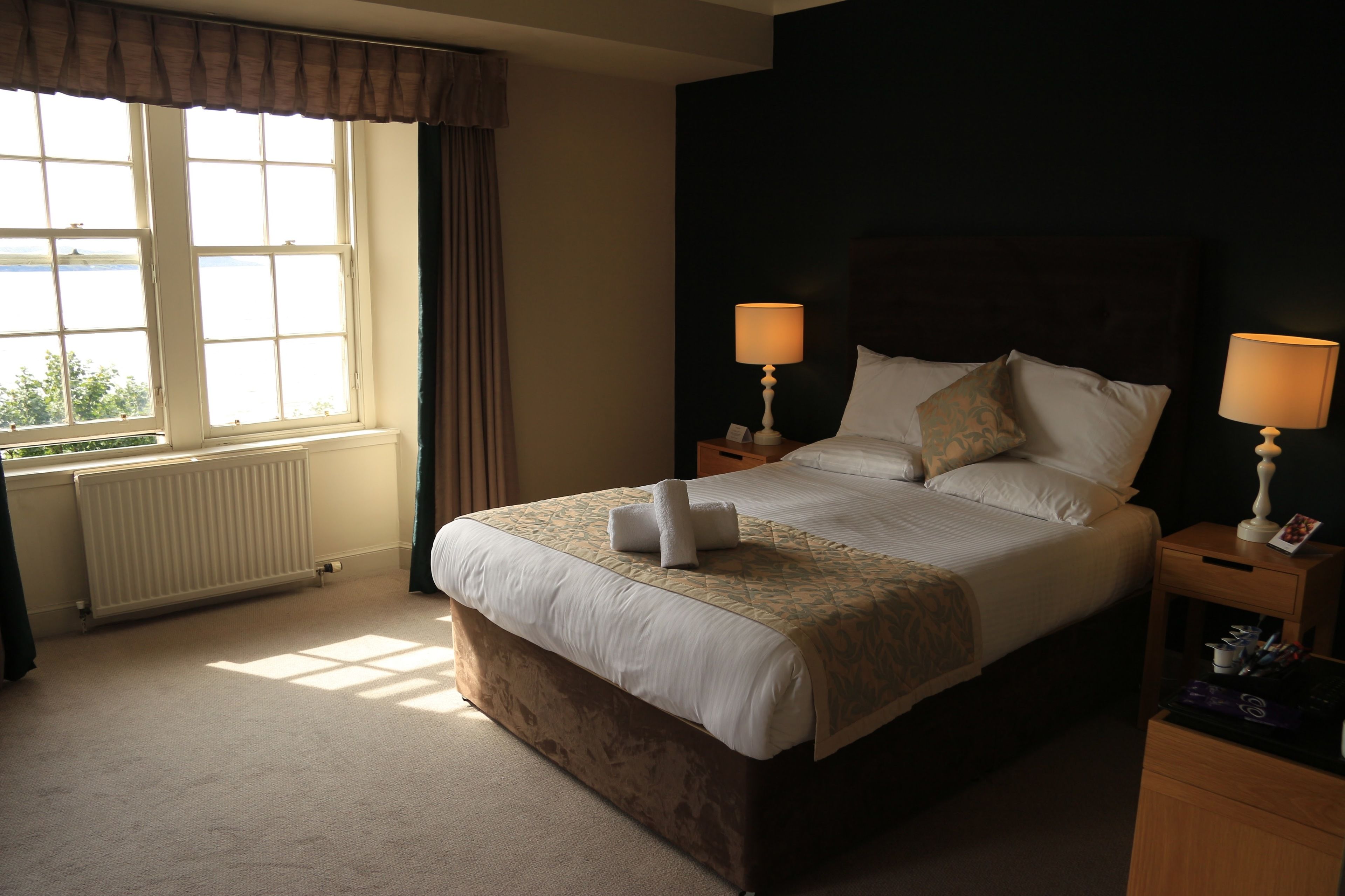 A neatly made bed with a white comforter and pillows, two bedside lamps, a wooden nightstand, and a view of the outdoors through a window with curtains.