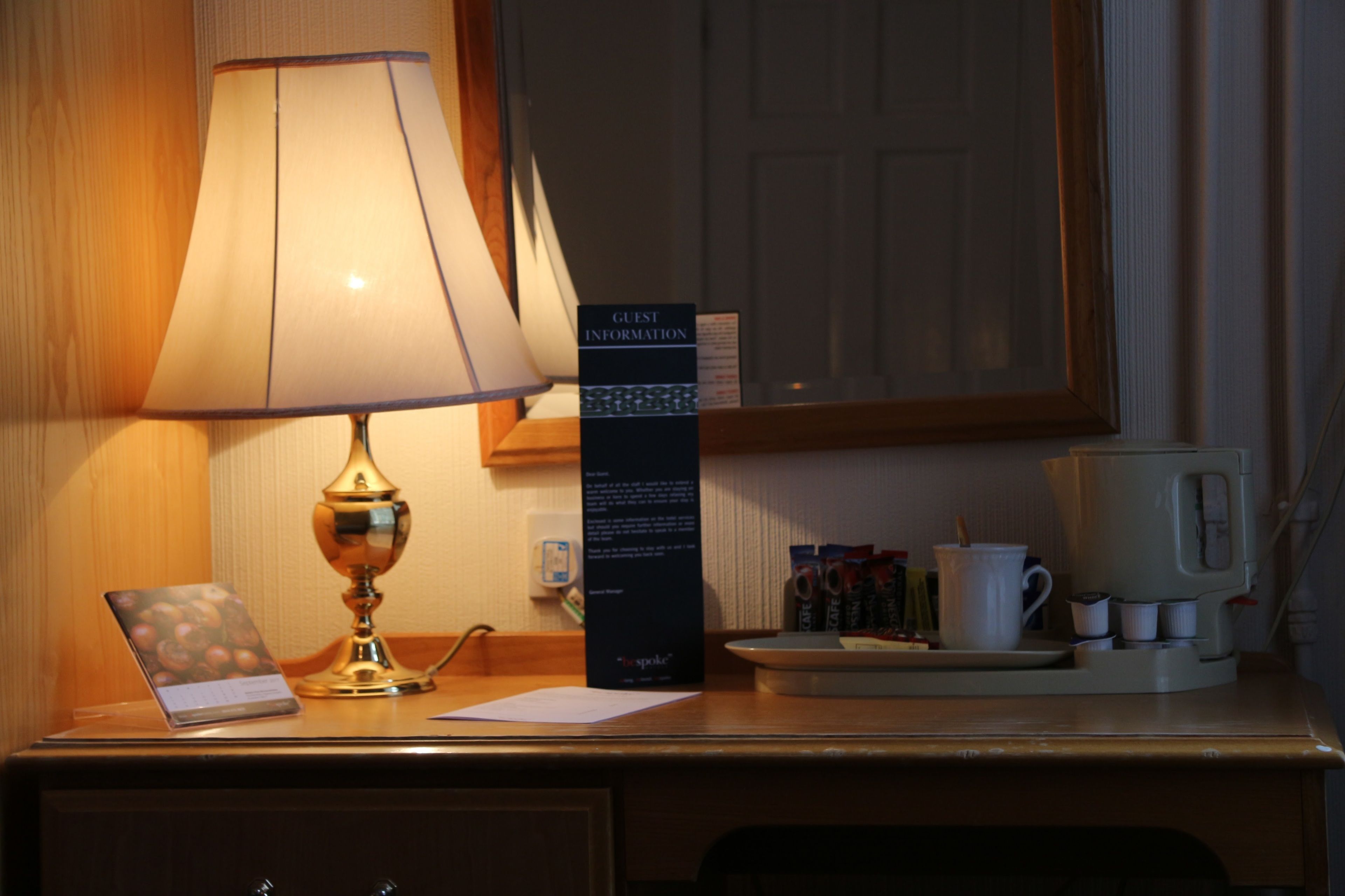 A hotel desk with a lamp, a sign for guest information, a laptop, a coffee maker, and a cup.
