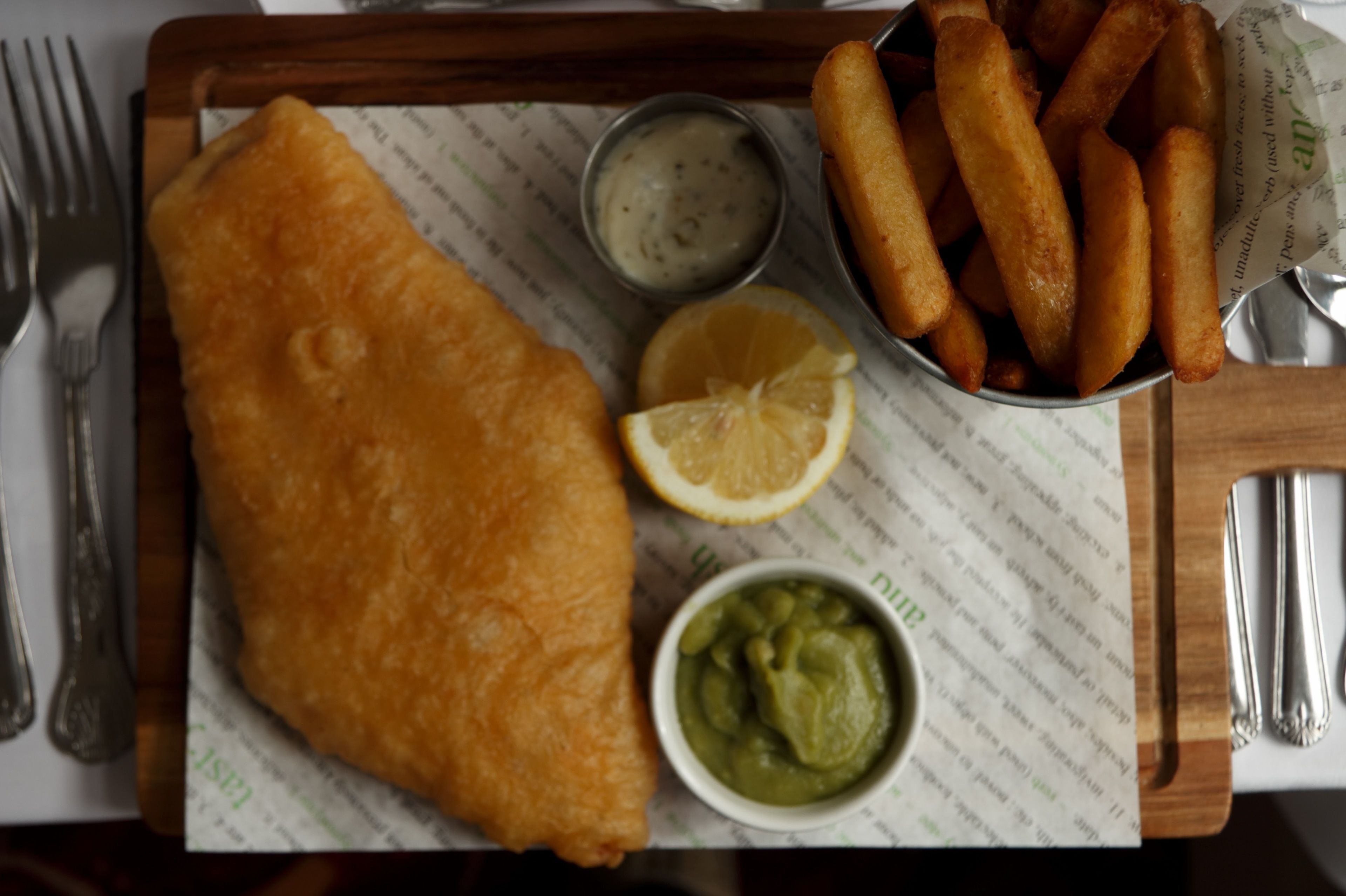 A meal of fish and chips with lemon and two dipping sauces on a wooden tray.