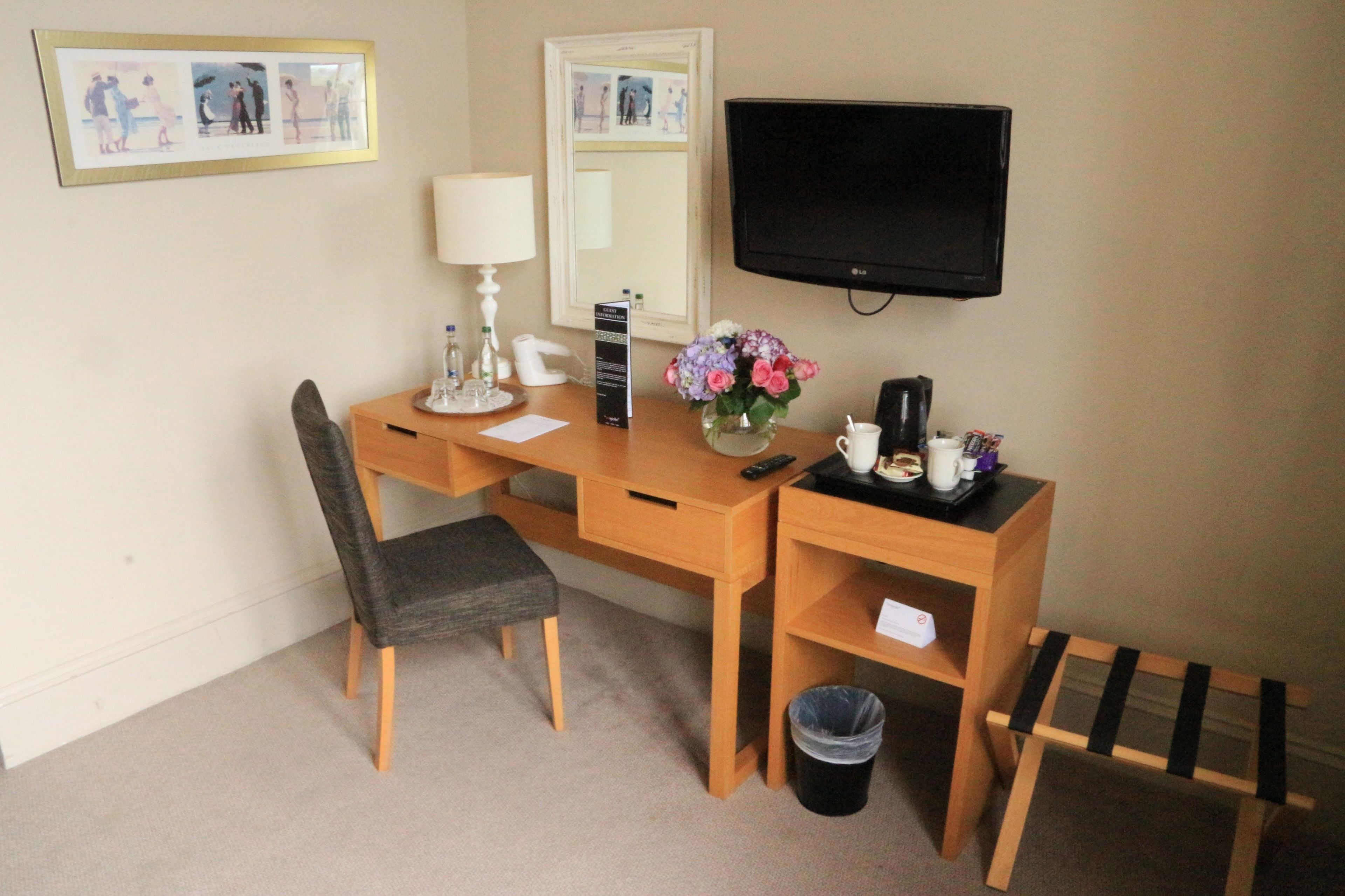 Ett hotellrum med ett skrivbord i trä, en stol, en tv, ett litet bord med en vas med blommor, en kaffebryggare och en papperskorg.