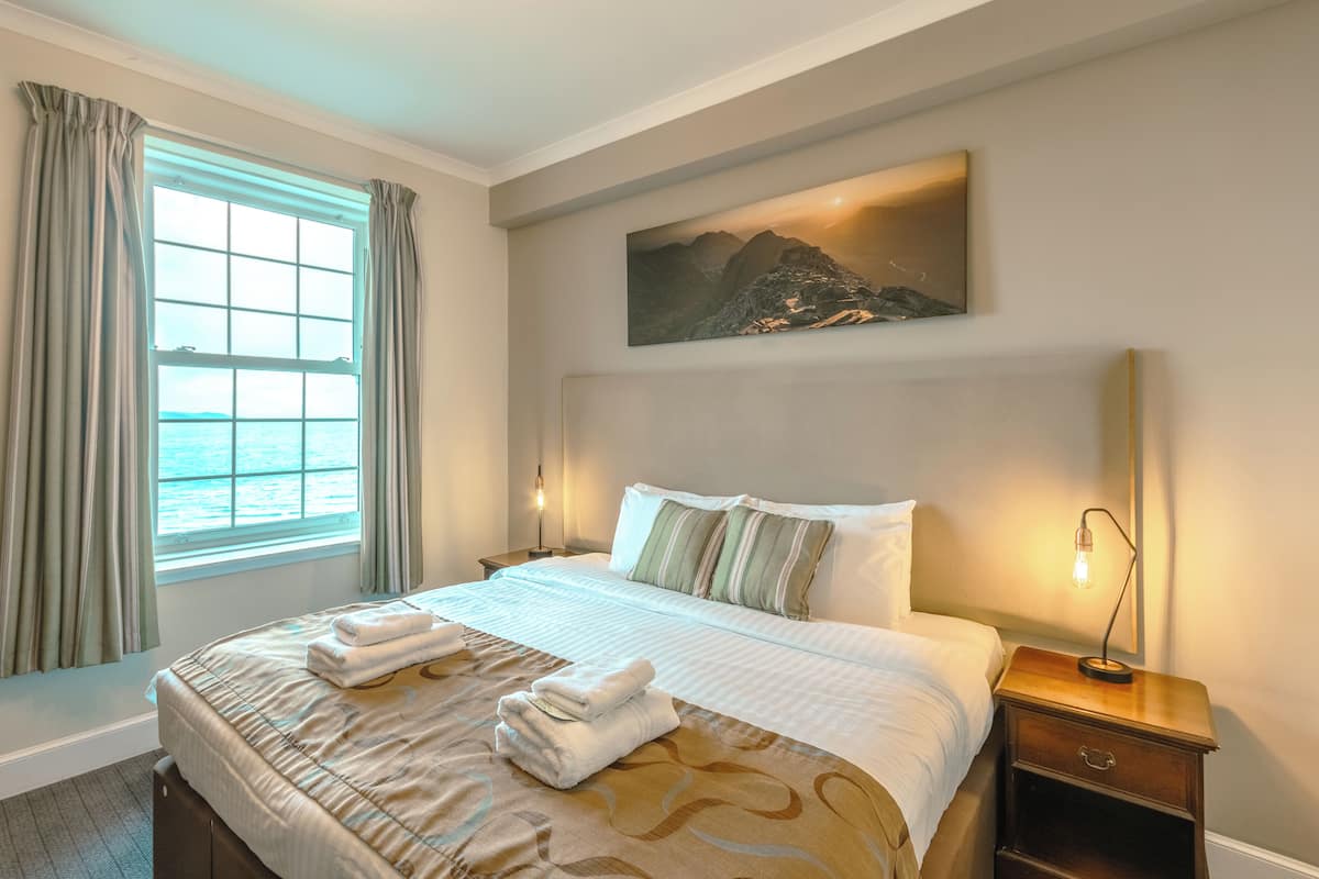 Een hotelkamer met een bed, nachtkastje, lamp, schilderij en een raam met gordijnen.