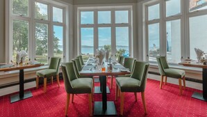 Un comedor con una mesa larga dispuesta para seis personas, rodeado de sillas verdes, ventanas amplias con vistas al mar y un suelo cubierto de alfombra roja.